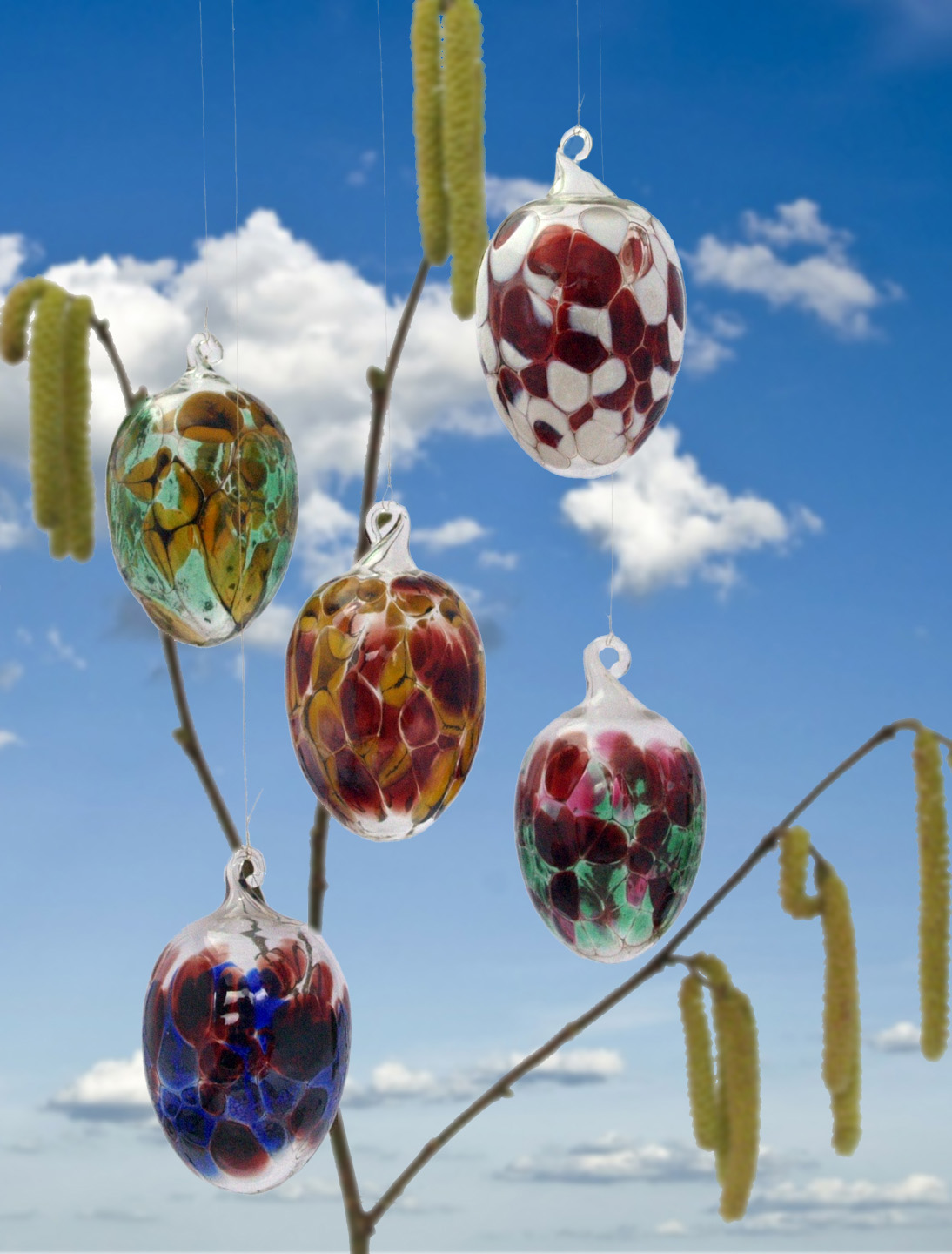 bunte Glas-Ostereier hängen an Ästen mit Weidenkätzchen vor blauem Himmel mit weißen Wolken